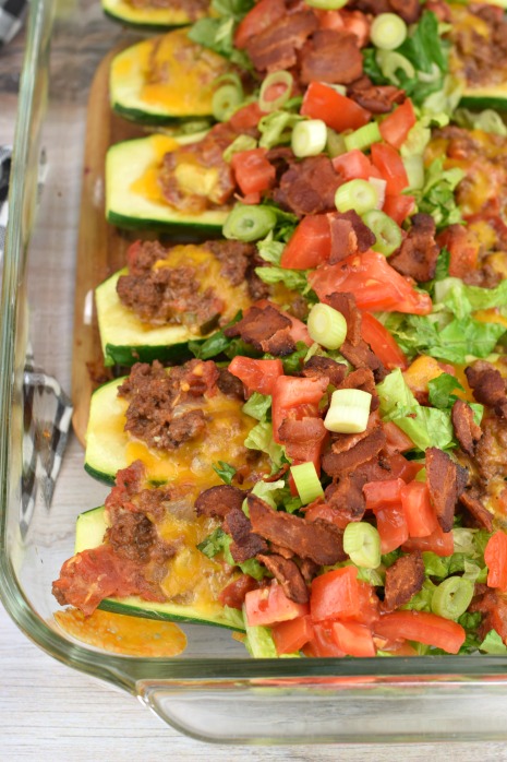 Zucchini boats in a 13x9 glass baking dish filled with bacon cheeseburger filling. Topped with cheese, bacon, lettuce, tomato and green onion.