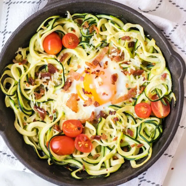 Cast iron skillet with zucchini nest topped with runny egg, bacon, and tomatoes.