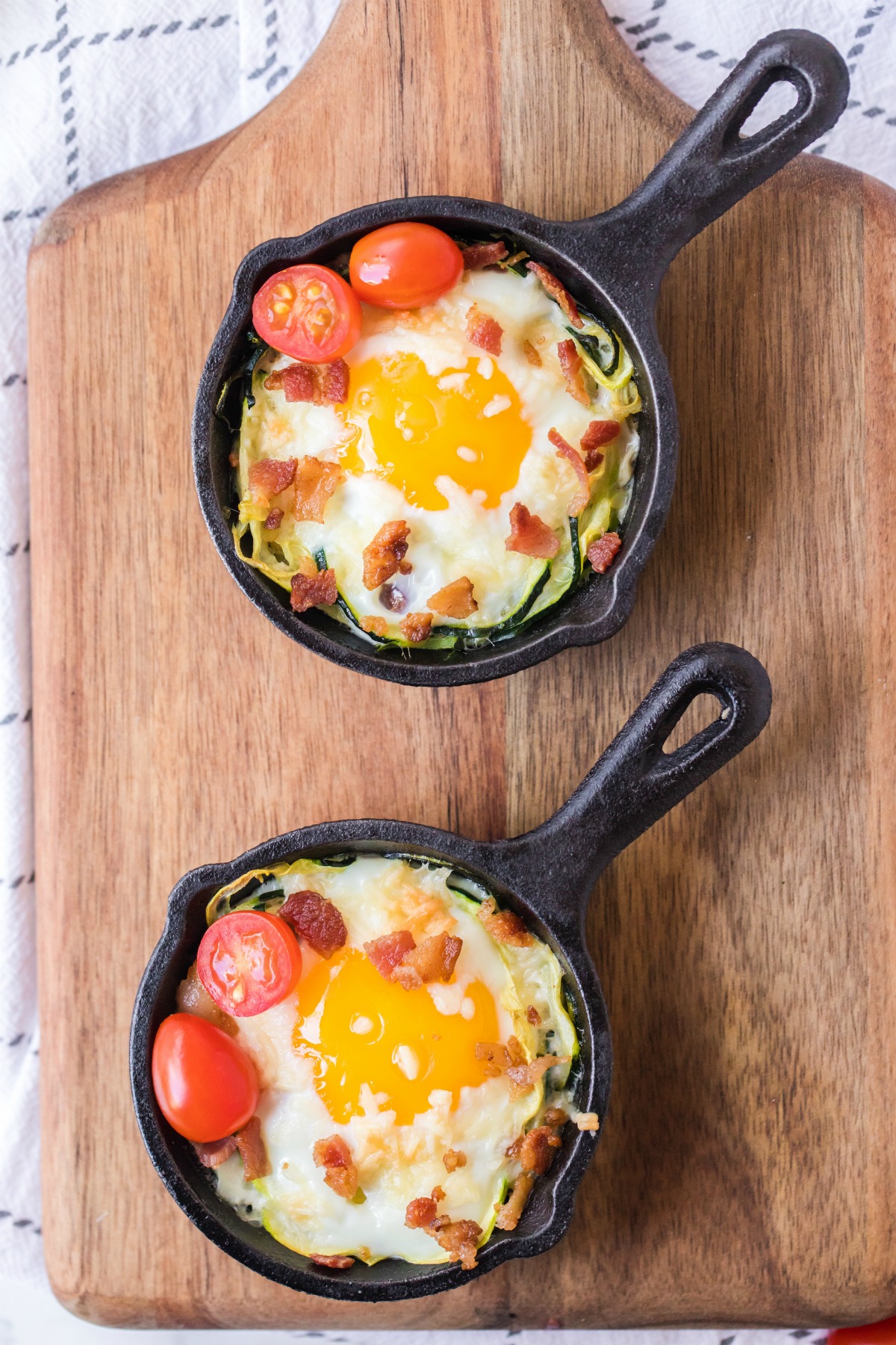 Two individual skillets with zucchini nest topped with runny egg and tomatoes.