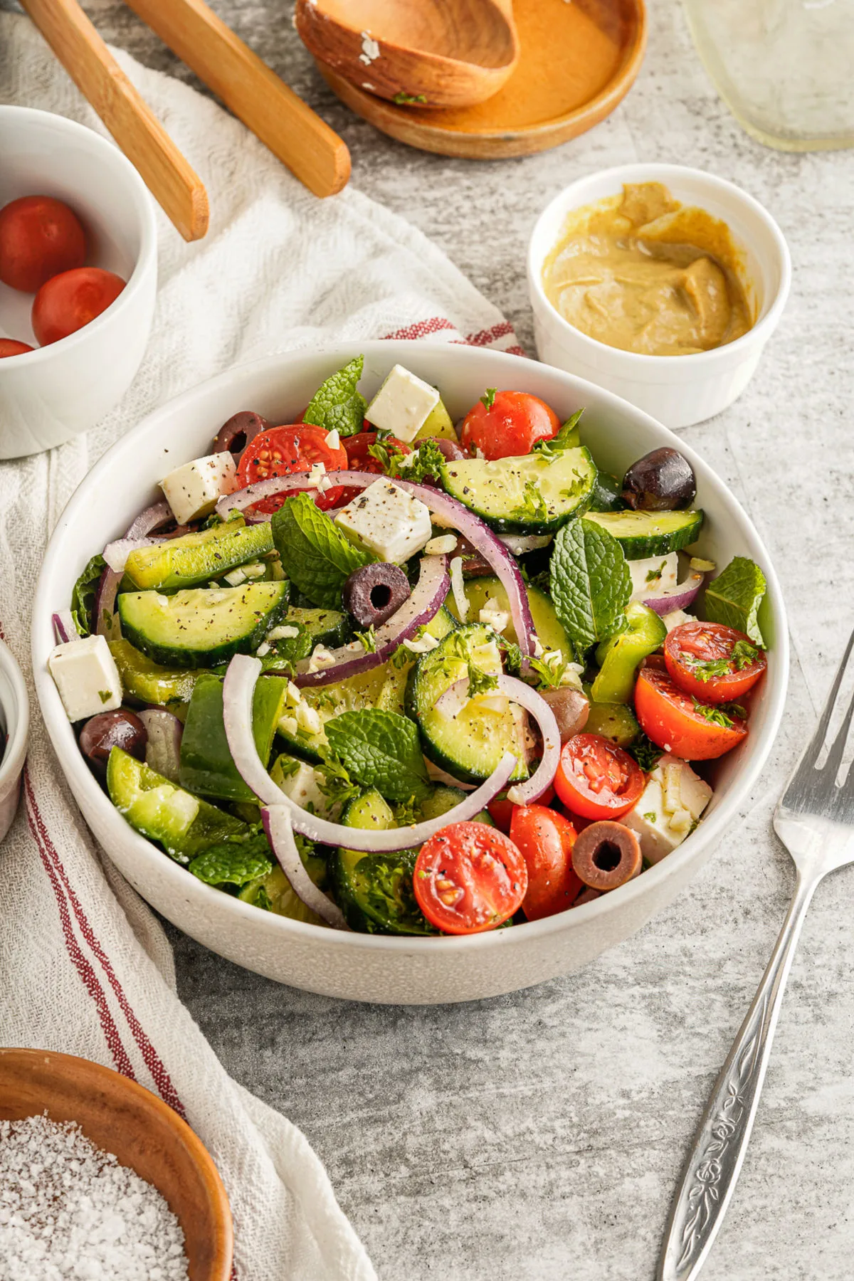 Greek salad in a white bowl with dressing ingredients in background.