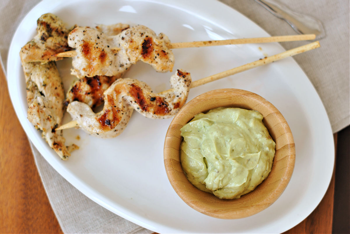 Chicken kebobs on white oval plate with wooden bowl of avocado sauce.