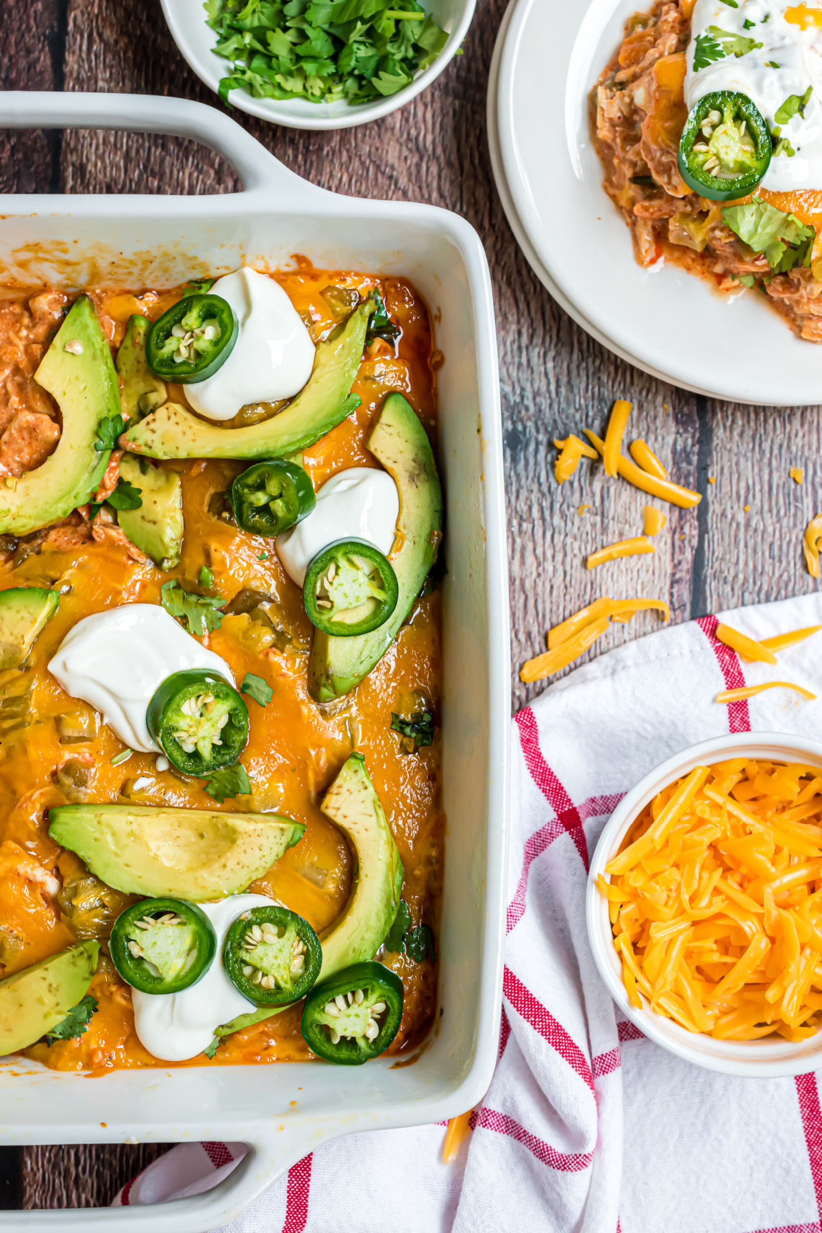 Chicken enchilada casserole in white baking dish served with avocado, cheese, sour cream, and jalapeno slices.